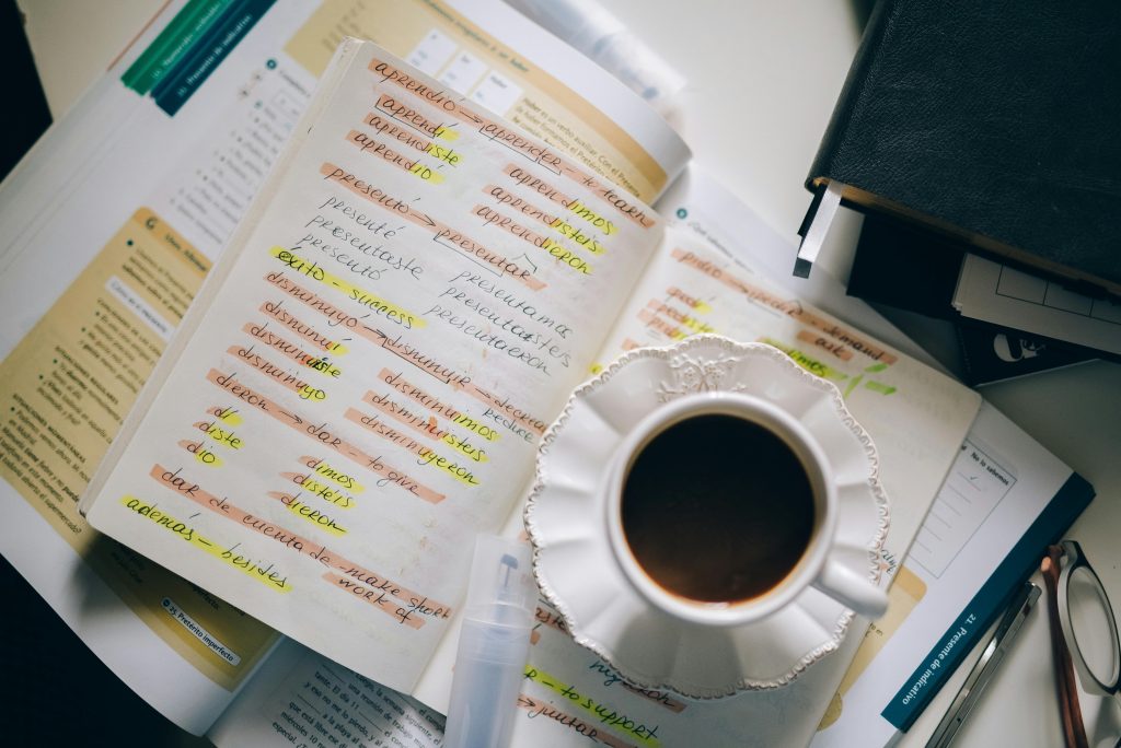 A picture of a desk with a Spanish grammar book, a notebook with verbs conjugations and a cup of black coffee.